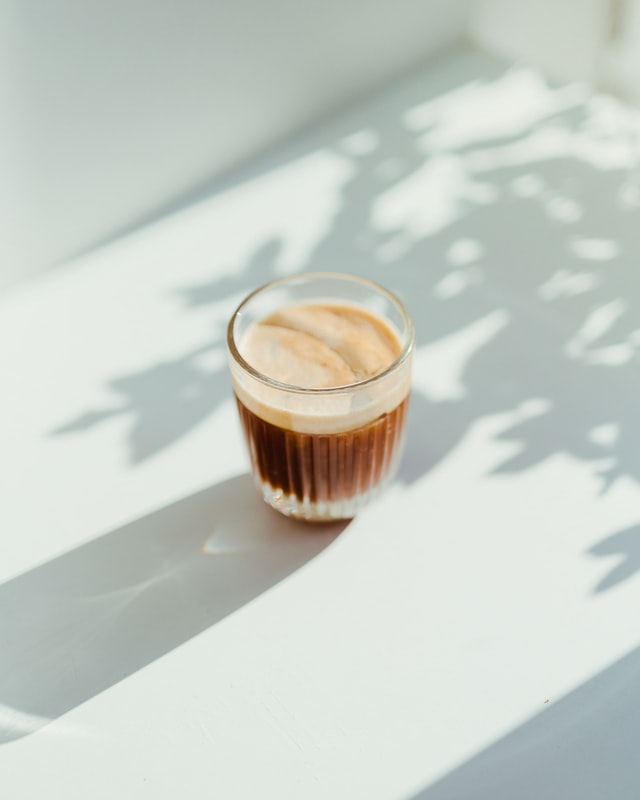 A glass of coffee on a white bench with the reflection of a plant around it