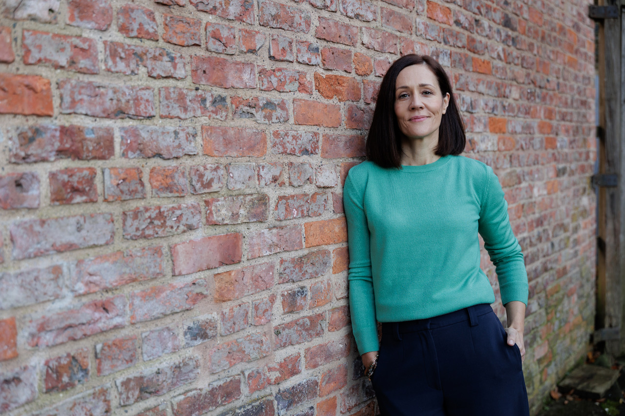 Background Sounds co-owner Mel Anderson wearing a green jumper and blue trousers. She has shoulder-length dark hair and is leaning against a brick wall