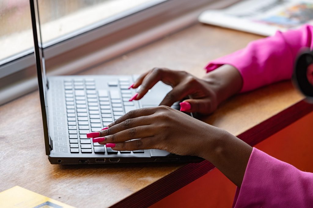 A black lady's hands with long pink nails typing on a laptop keyboard