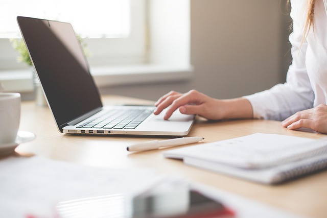 Open laptop on a desk with a woman's hand on the keyboard, open notebook and pen in the foreground