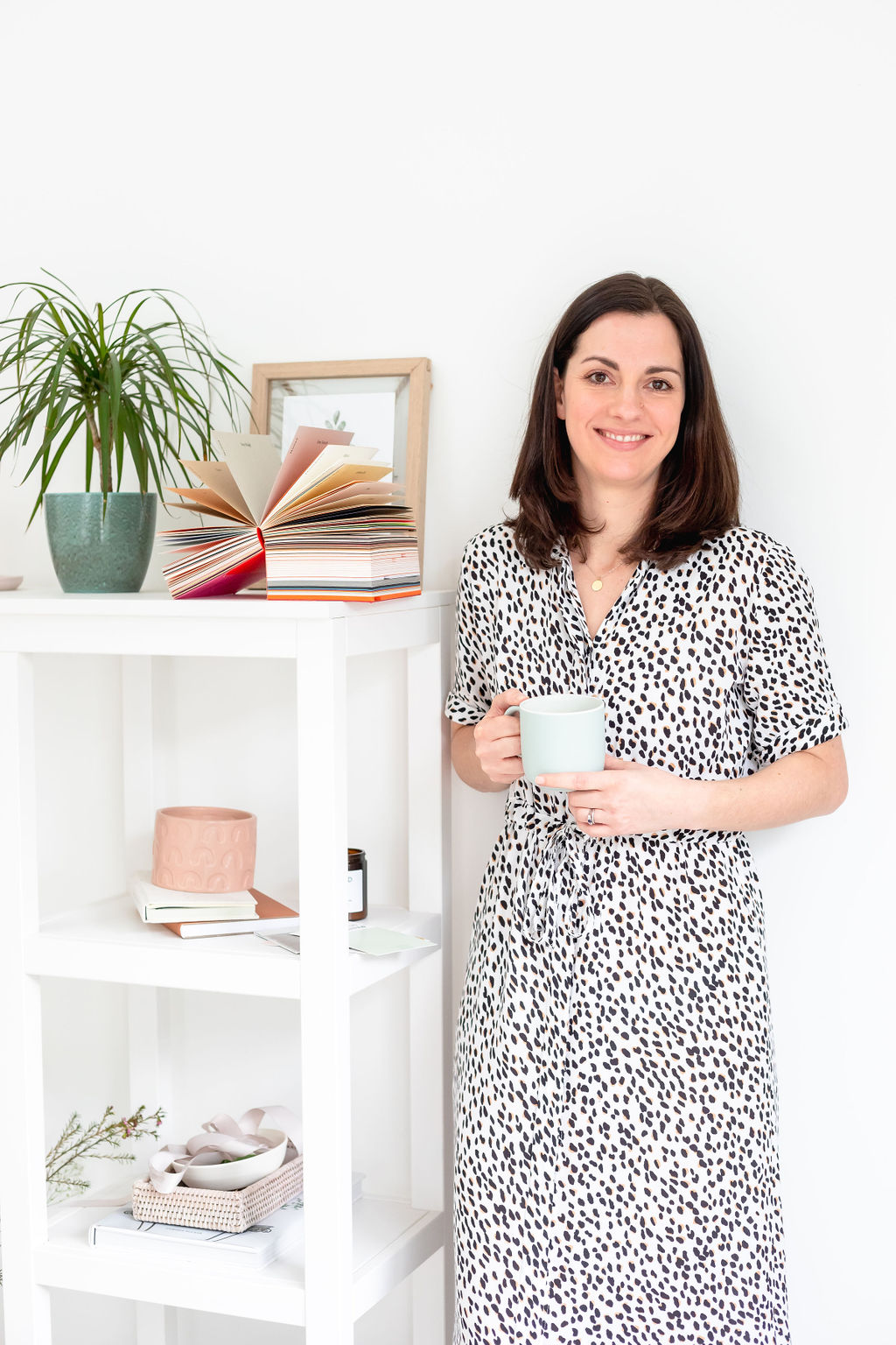 Jane from JM Creative wearing a black and white print dress, holding a cup and standing beside a white shelf unit